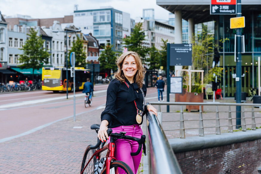 Belle de Gast staand, met een racefiets aan de hand, op het Vredenburg in Utrecht. Ze heeft haar burgemeestersketting om.